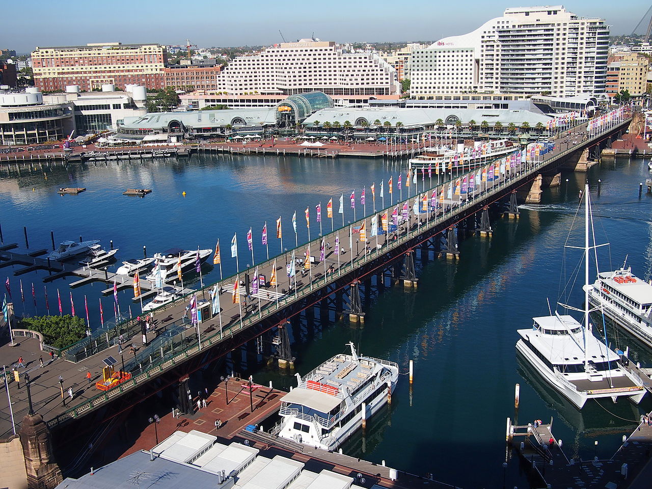 The Pyrmont Bridge viewed from the north-east after the removal of the monorail track. Opened in 1902, the Bridge connects Pyrmont and the city as a pedestrian route. The 369m bridge consists of 14 spans, 12 of which are 25m long fixed timber trusses, with the remaining two being steel swing spans. The Bridge features 72 Allan timber trusses spaced approximately three meters apart. (Photo Credit: Nick-D shared by Wikimedia using a  Creative Commons Attribution-Share Alike 3.0 Unported license)